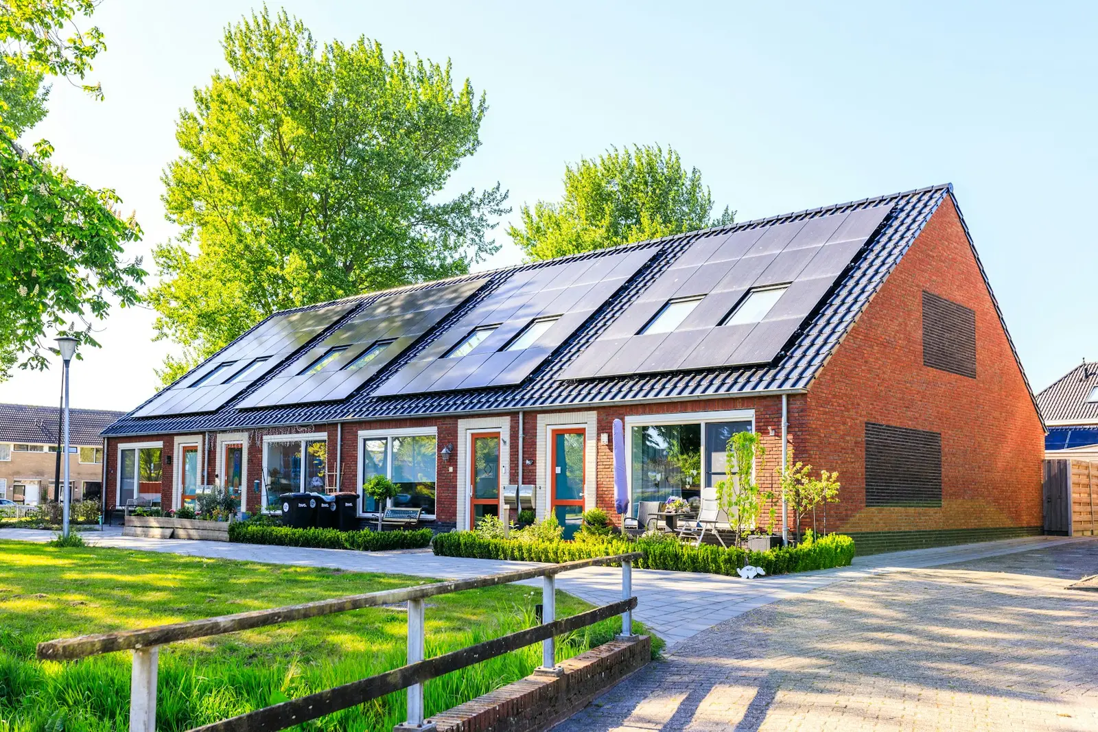 Een rij nieuwe bakstenen rijtjeshuizen met zonnepanelen op het hele dak, in de zon, met een grasstrook en een hek ervoor